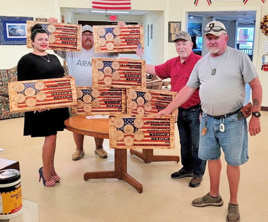 A group of people holding wooden plaques engraved with American flags and military logos, standing together in a room with patriotic decorations.