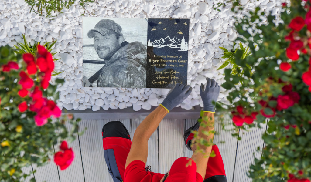 Person arranging a laser-engraved home garden memorial plaque with a portrait and inscription, surrounded by white stones and red roses.